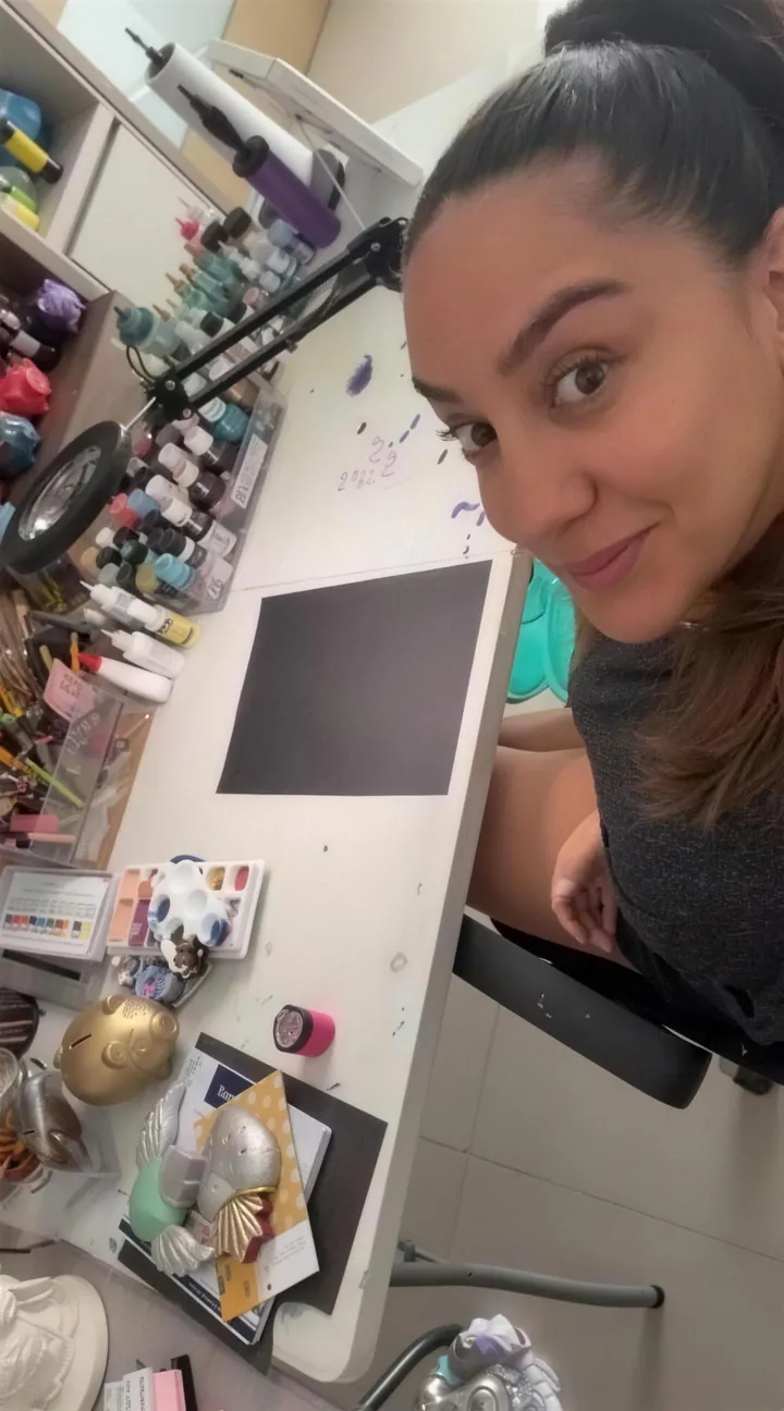 Stephie sonríe sentada en su mesa de trabajo, frente a una hoja de papel negro, rodeada de pinturas.