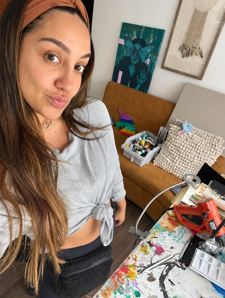Stephie mira a la cámara en su casa; detrás, en la pared, uno de sus cuadros, y abajo su mesa llena de pinturas y pinceles.